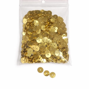 May include: A clear plastic bag filled with numerous gold-colored sequins. The sequins are round with a hole in the center. A few sequins are scattered outside the bag. The bag has a red zipper closure.