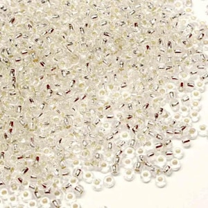 May include: A large quantity of small, clear glass seed beads with silver accents. The beads are scattered on a white surface.
