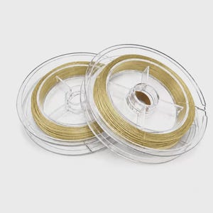 Puede incluir: Dos bobinas de plástico transparente enrolladas con alambre metálico de color dorado. Las bobinas presentan un diseño circular con radios internos y un eje central, que sostiene una longitud continua de alambre fino enrollado alrededor.