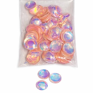 May include: A clear plastic bag containing iridescent, circular sequins. The sequins shift in colour from pink to purple. A few sequins are scattered outside the bag. These sequins are suitable for craft projects and embellishments.