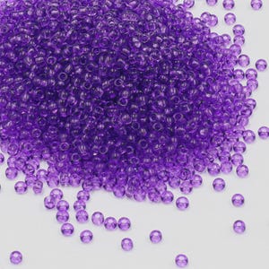 Könnte beinhalten: Ein Haufen kleiner, durchscheinender, violetter Glasperlen. Die Perlen sind auf einer weißen Oberfläche verstreut, wobei sich die meisten zusammenballen. Die Perlen sind rund mit einem Loch in der Mitte, geeignet für Schmuckherstellung oder Bastelarbeiten.