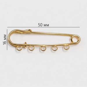 May include: A gold-colored safety pin with five small loops for attaching charms or beads. The pin measures 50 mm long and 16 mm wide.