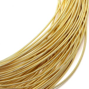May include: A coiled length of metallic gold-colored wire featuring a tightly wound, ribbed, or spring-like texture. The wire is arranged in multiple overlapping loops, creating a dense, uniform pattern of reflective, cylindrical strands.