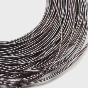 May include: A coil of dark metallic wire featuring a continuous, tightly wound spiral texture. The wire has a smooth, reflective surface with a deep gunmetal gray finish, arranged in multiple overlapping circular loops that create a dense pattern.