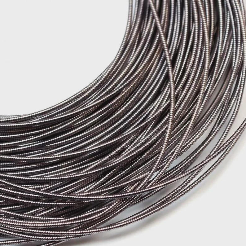 May include: A coil of dark metallic wire featuring a continuous, tightly wound spiral texture. The wire has a smooth, reflective surface with a deep gunmetal gray finish, arranged in multiple overlapping circular loops that create a dense pattern.
