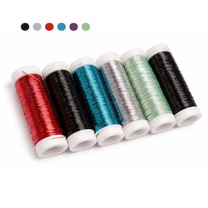 Könnte beinhalten: Sechs Spulen Metallic-Garn in verschiedenen Farben: Rot, Schwarz, Türkis, Silber, Mintgrün und Schwarz. Jede Spule hat eine weiße Basis. Die Fäden sind eng gewickelt und weisen eine glänzende, reflektierende Qualität auf, ideal für Bastel- und Stickprojekte.