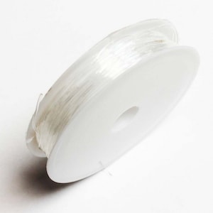 Könnte beinhalten: Eine Spule mit klarem elastischem Faden, die um eine weiße Kunststoffspule gewickelt ist.