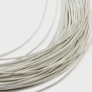 May include: A coil of silver-colored metallic wire featuring a fine, textured, coiled surface. The wire is arranged in multiple concentric loops, creating a dense, circular pattern with a consistent, reflective, and bright metallic finish throughout.