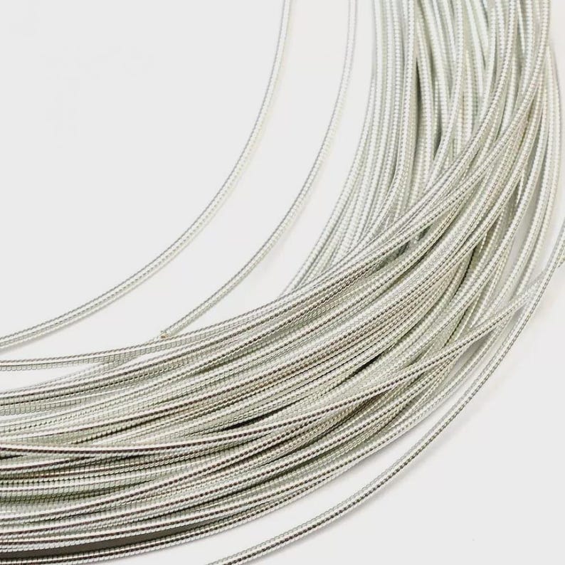 May include: A coil of silver-colored metallic wire featuring a fine, textured, coiled surface. The wire is arranged in multiple concentric loops, creating a dense, circular pattern with a consistent, reflective, and bright metallic finish throughout.