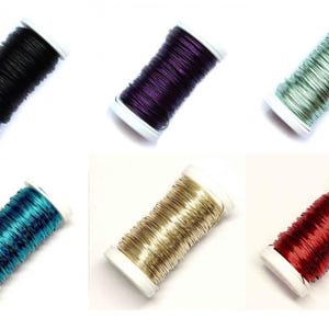 Op de afbeelding: Zes spoelen draad in verschillende kleuren: zwart, paars, groen, blauwgroen, goud en rood. Elke spoel is gewikkeld rond een witte plastic kern, ideaal voor het maken van sieraden of knutselen.