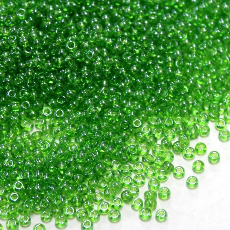 May include: A large quantity of small, round, green glass beads. The beads have a hole in the center and are scattered on a white surface.