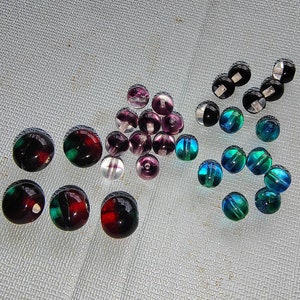 May include: Assortment of colorful glass beads in various shades of purple, green, and black. The beads are round and have a smooth, shiny finish.