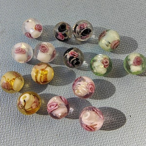May include: A collection of glass beads with floral designs. The beads are round and feature various colors, including pink, gold, black, and white. Some beads have a rose pattern, while others have gold or silver accents. The beads are scattered on a light blue surface.