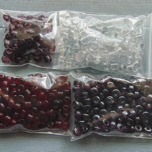 May include: Four clear plastic bags filled with beads. The beads are in various colors, including clear, dark red, and dark purple. The beads are small and cylindrical, suitable for jewelry making or crafts.
