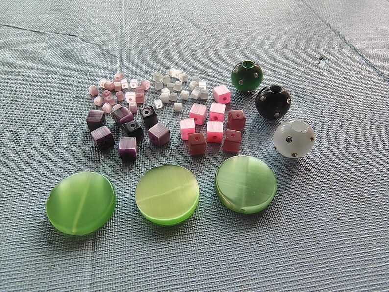 May include: Assortment of colorful beads, including green, black, white, pink, and purple. The beads are made of glass and plastic and are in various shapes, including round, square, and cube. There are also three large green and white beads.