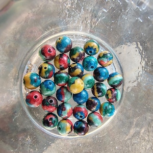 May include: A collection of colorful, round beads with a marbled effect. The beads are primarily blue, red, yellow, and green, and are contained within a clear glass bowl. Each bead has a small hole.