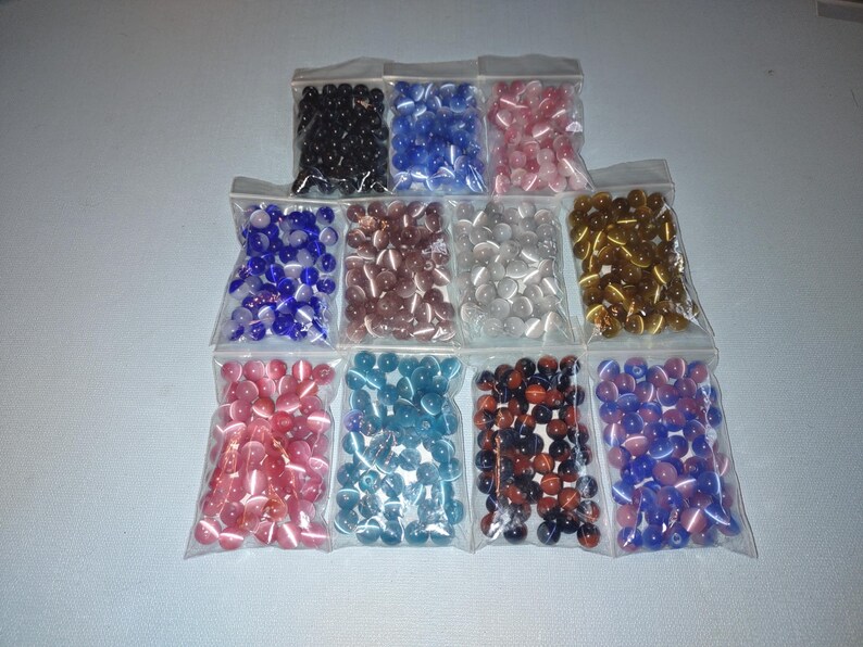 K&ouml;nnte beinhalten: Sortiment kleiner, runder Glasperlen in verschiedenen Farben, darunter Blau, Rosa, Wei&szlig;, Gold, Rot und Schwarz. Die Perlen sind in durchsichtigen Plastikt&uuml;ten verpackt.