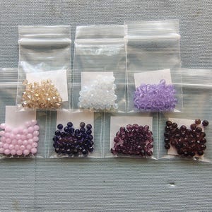 May include: Assortment of colorful beads in small, clear plastic bags. The beads are in various colors, including gold, white, purple, pink, dark blue, and brown. Each bag contains a different color of beads, perfect for jewelry making.