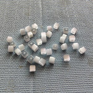 May include: A collection of white and gray square beads with a hole in the center. The beads are scattered on a blue textured surface.