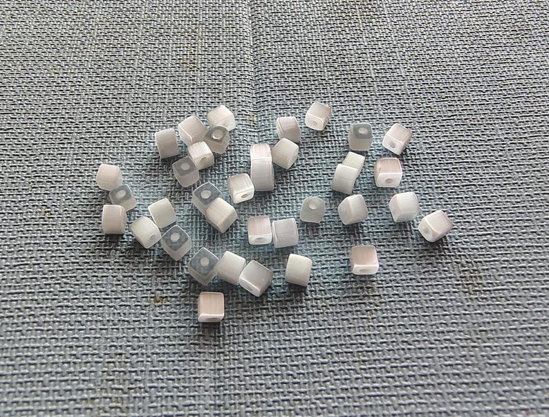 May include: A collection of white and gray square beads with a hole in the center. The beads are scattered on a blue textured surface.