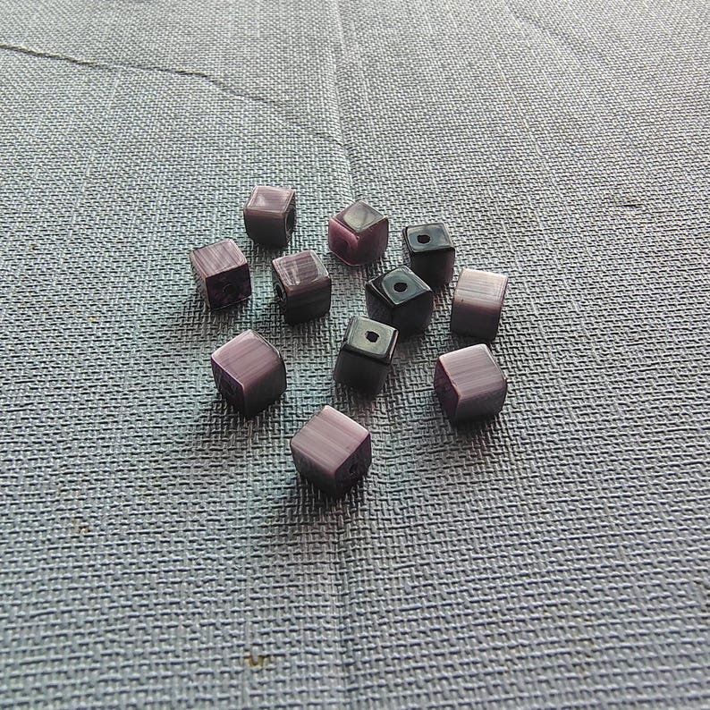 May include: A collection of purple and black square-shaped beads. The beads have a marbled effect and are scattered on a gray textured surface.