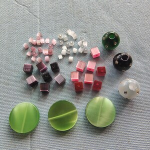 May include: Assortment of colorful glass beads in various shapes and sizes, including round, square, and oval. The beads are in shades of pink, purple, green, and white. There are also three larger round beads with a textured surface.