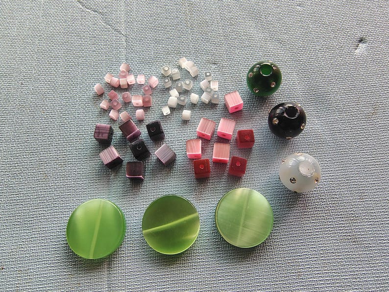 May include: Assortment of colorful glass beads in various shapes and sizes, including round, square, and oval. The beads are in shades of pink, purple, green, and white. There are also three larger round beads with a textured surface.