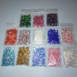 K&ouml;nnte beinhalten: Sortiment kleiner Glasperlen in verschiedenen Farben, darunter Blau, Gr&uuml;n, Rosa, Wei&szlig; und Gold, verpackt in durchsichtigen Plastikt&uuml;ten.