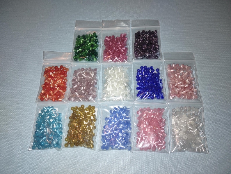 K&ouml;nnte beinhalten: Sortiment kleiner Glasperlen in verschiedenen Farben, darunter Blau, Gr&uuml;n, Rosa, Wei&szlig; und Gold, verpackt in durchsichtigen Plastikt&uuml;ten.
