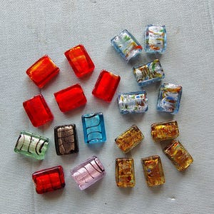 May include: A collection of colorful, rectangular glass beads in various colors, including red, blue, gold, and green. The beads have a textured, translucent appearance, with some featuring internal patterns.