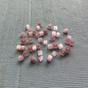 May include: A group of small, square, translucent pink beads with a white iridescent sheen.