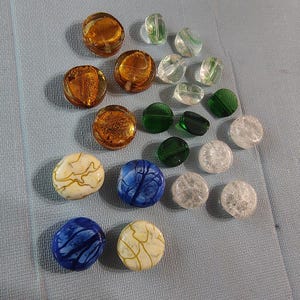 May include: A collection of colorful beads in various shapes and sizes. The beads include gold-flecked, clear, green, white, and blue designs. Some are round, while others are more irregular in shape. The beads are scattered on a light blue surface.