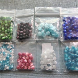 May include: Assortment of colorful glass beads in various shapes and sizes, packaged in clear plastic bags. The beads are in shades of green, purple, blue, pink, and white.