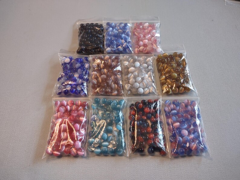 K&ouml;nnte beinhalten: Zehn kleine Beutel mit Glasperlen in verschiedenen Farben, darunter Blau, Rosa, Braun, Gold, Rot und T&uuml;rkis. Die Perlen sind alle rund und haben ein wei&szlig;es Spiralmuster.