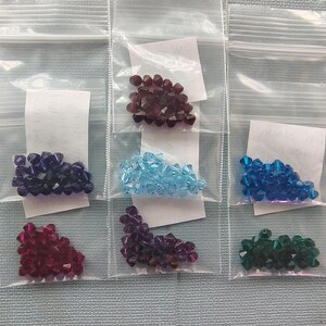 May include: Six small plastic bags each containing a pile of faceted beads in various colors. Colors include purple, red, blue, and green. Each bag has a small white paper insert.