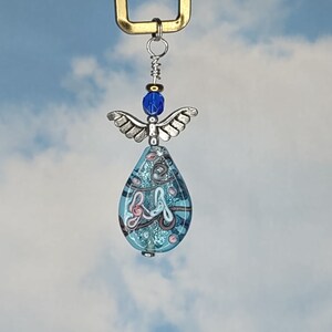 May include: A blue glass angel ornament with silver wings and a blue and pink swirl design. The ornament is hanging from a gold hook.