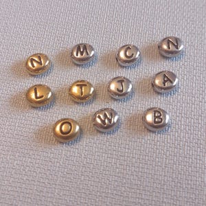 May include: A collection of silver and gold letter beads, each bead features a single letter of the alphabet. The letters are: N, M, C, A, J, B, L, T, O, W.