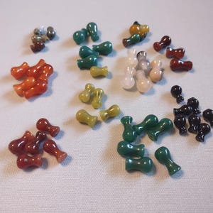 May include: Assortment of small, colorful glass beads in various shapes and colors, including green, red, black, white, and yellow. The beads are arranged in a circular pattern on a white surface.