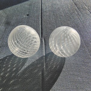 May include: Two clear glass spheres with a white, striped pattern. The spheres are sitting on a gray textured surface.