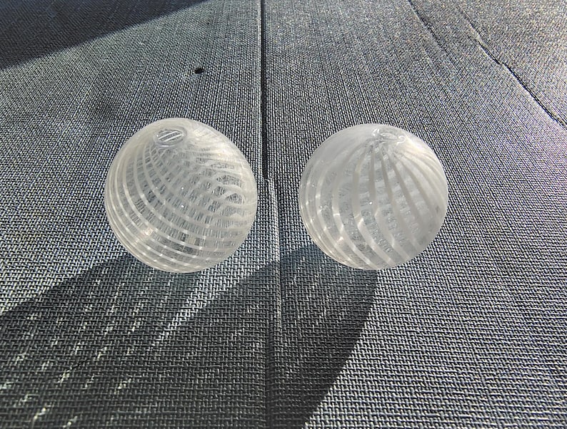 May include: Two clear glass spheres with a white, striped pattern. The spheres are sitting on a gray textured surface.