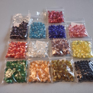 K&ouml;nnte beinhalten: Sortiment kleiner, ovaler Glasperlen in verschiedenen Farben, verpackt in durchsichtigen Plastikt&uuml;ten. Die Farben umfassen Rot, Blau, Gr&uuml;n, Gelb, Wei&szlig; und Schwarz.