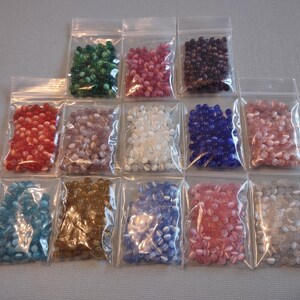 K&ouml;nnte beinhalten: Sortiment von 12 kleinen Beuteln mit Glasperlen in verschiedenen Farben und Mustern. Die Perlen sind oval geformt und haben eine gl&auml;nzende Oberfl&auml;che.