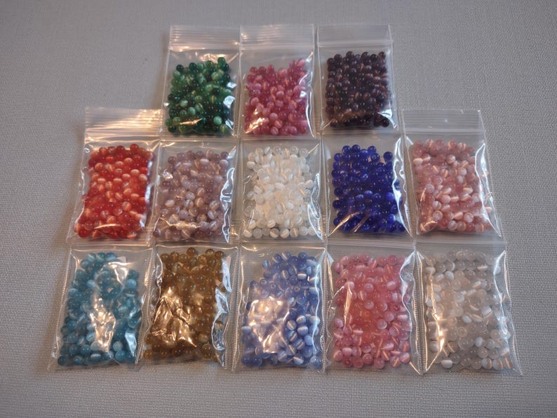 K&ouml;nnte beinhalten: Sortiment von 12 kleinen Beuteln mit Glasperlen in verschiedenen Farben und Mustern. Die Perlen sind oval geformt und haben eine gl&auml;nzende Oberfl&auml;che.