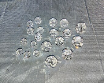 May include: A collection of clear glass beads with faceted surfaces. The beads are arranged in a heart shape on a gray textured surface.