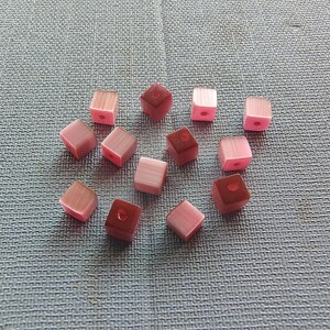 May include: A collection of 13 pink and red square beads with a hole in the center. The beads are made of glass and have a translucent finish.