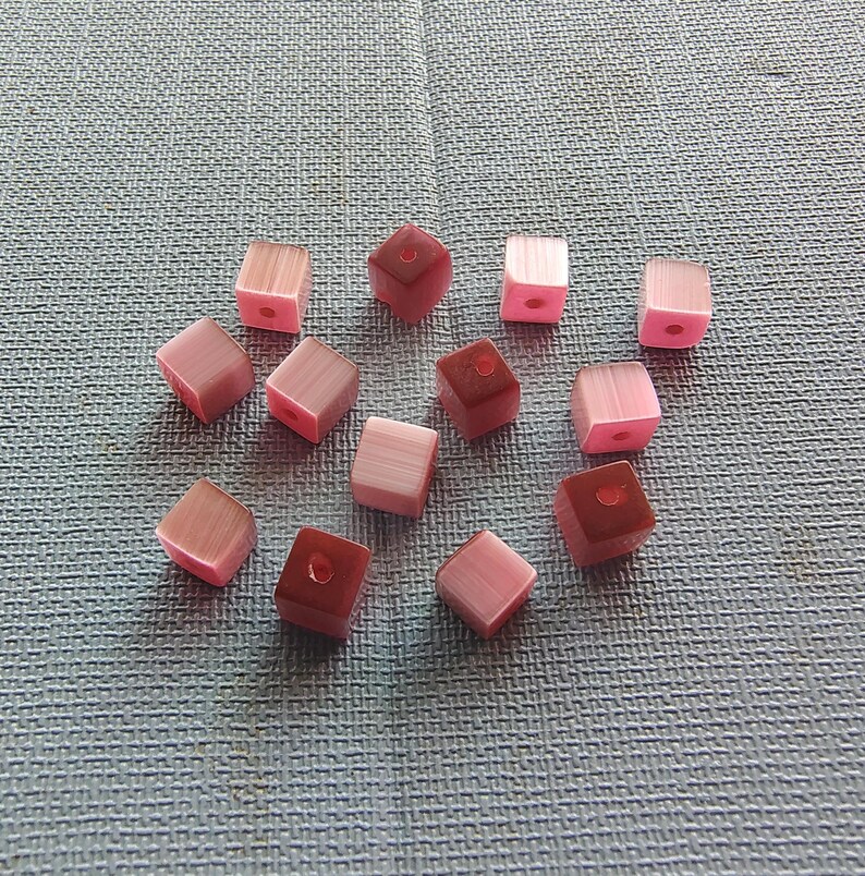 May include: A collection of 13 pink and red square beads with a hole in the center. The beads are made of glass and have a translucent finish.