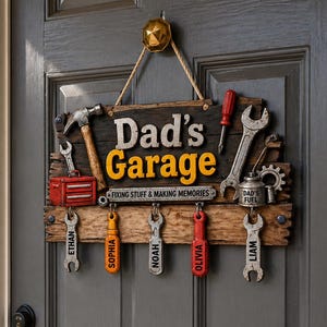 May include: A wooden sign for a garage, with the words "Dad's Garage" in yellow and white. It features tool-themed keychains with names like Ethan, Sophia, and Liam. The sign hangs on a gray door.