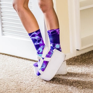 May include: A pair of white platform sandals with a tie-dye pattern on the straps. The sandals are worn with purple and blue tie-dye socks.