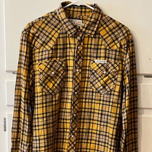 Puede incluir: Una camisa de manga larga a cuadros amarillos y negros con cuello puntiagudo y dos bolsillos en el pecho.