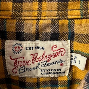 Puede incluir: Una camisa de cuadros amarilla, negra y azul con una etiqueta blanca que dice "True Religion Brand Jeans" y "EST 1956" en letras rojas. La etiqueta tambi&eacute;n dice "Styled in California" y "Made in China".